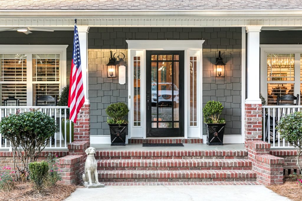 Front door entrance to a large two story blue gray house with wood and vinyl siding and a large American flag.
